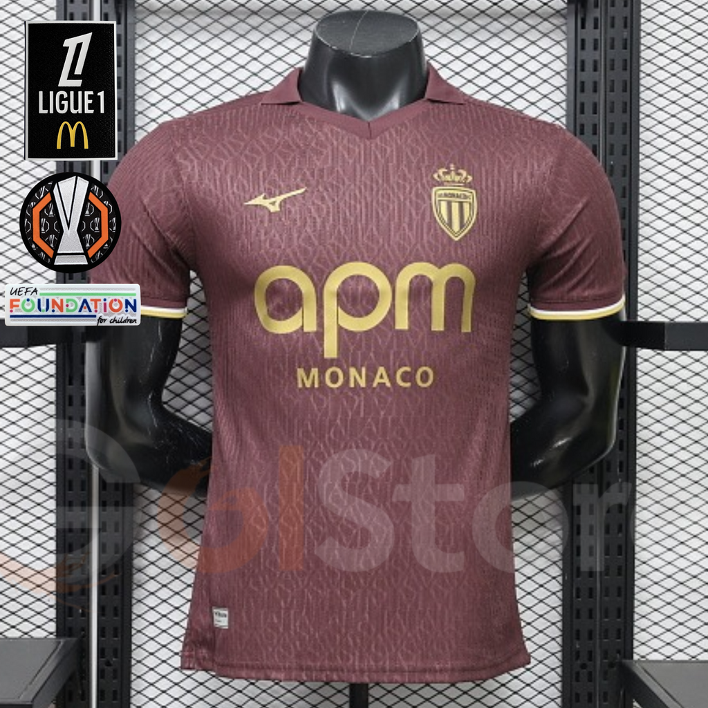 AS Monaco burgundy short-sleeve jersey with tonal pattern, gold crest and APM Monaco sponsor, front view alongside Ligue 1 and Europa League graphics.
