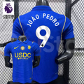 Front and back views of a blue Chelsea short-sleeve jersey with yellow details, white João Pedro number 9 print and a gold chest badge, alongside a Premier League graphic.