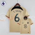 Front and back views of a pale gold Chelsea FC 2022–2023 short-sleeve third jersey with dark T. Silva #6 printing, a black upper back panel and black and orange sleeve trim.