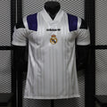 Front view of a white Real Madrid short-sleeve jersey with tonal vertical stripes, purple and black shoulder panels, a white collar, black adidas logo and centered club crest.