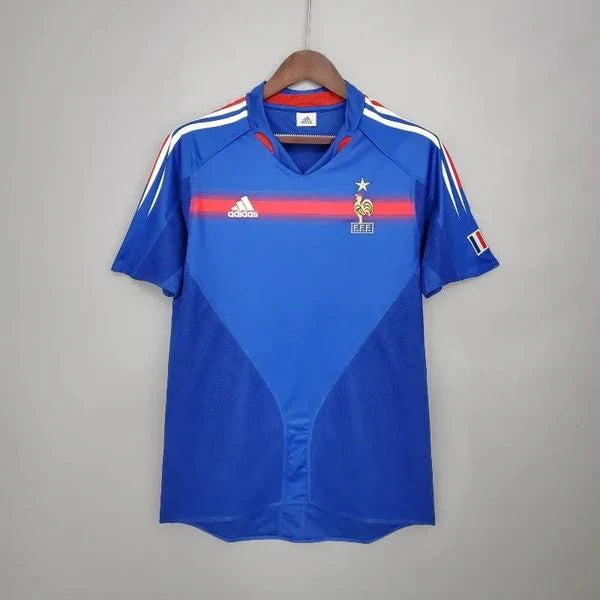 Front view of a France 2004 retro blue short-sleeve home jersey with a red chest stripe, white shoulder stripes, national team crest and French flag sleeve detail.