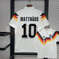  Front and back views of a white Germany 1990 retro short-sleeve home jersey with black, red and gold stripes and black Matthäus number 10 print.