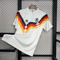 Front view of a white Germany 1990 retro short-sleeve home jersey with black, red and gold geometric chest stripes and the German football crest.