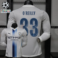 Front and back views of a white Manchester City long-sleeve jersey with blue central stripes, blue O’Reilly number 33 print and Etihad Airways sponsor, alongside UCL graphics.
