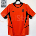 Netherlands orange short-sleeve retro jersey with black collar, black side panels and KNVB lion crest, alongside a 2002 World Cup patch graphic.
